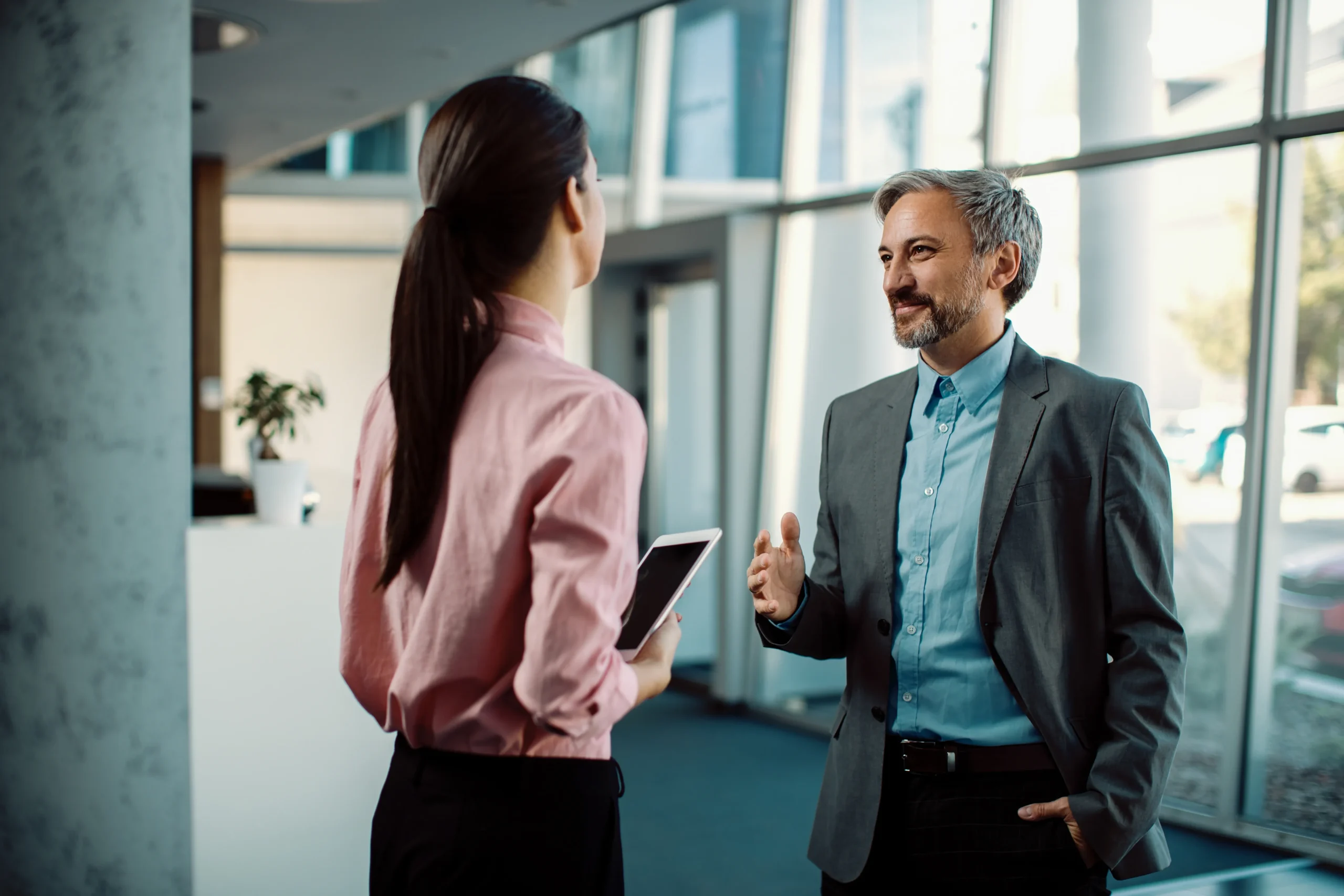 Empresário sorridente conversa com colega segurando tablet em escritório corporativo moderno.