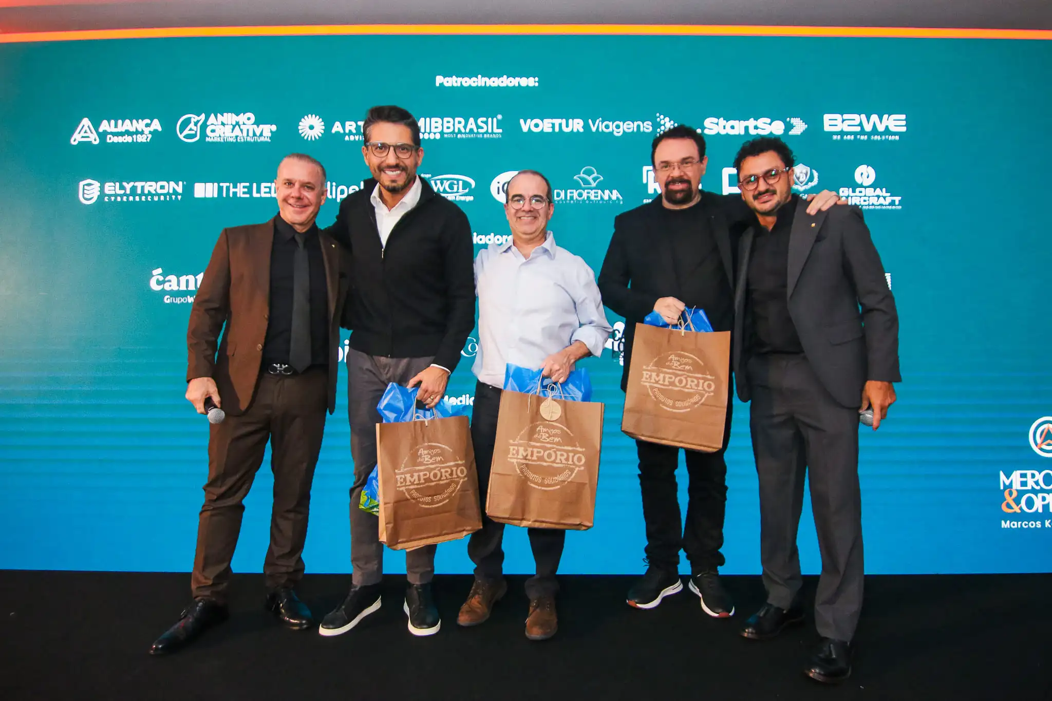 Cinco homens posam sorrindo em evento corporativo segurando sacolas, com painel de patrocinadores ao fundo.