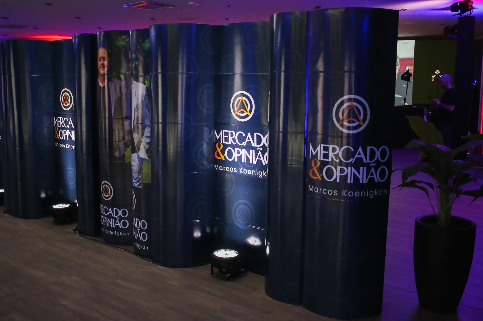 Painéis do programa Mercado & Opinião com Marcos Koenigkan em evento corporativo com iluminação roxa.