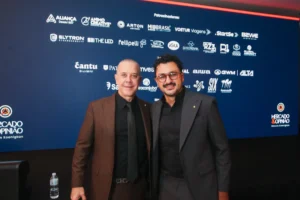 Dois homens de terno sorriem posando para foto no evento Mercado & Opinião, diante de painel azul com logotipos dos patrocinadores.