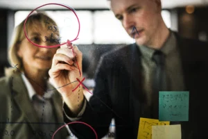 Homem e mulher de negócios planejando estratégia em parede de vidro com gráficos e anotações.
