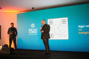 Dois homens de terno no palco do evento Mercado & Opinião, um falando ao microfone, com painel azul ao fundo.