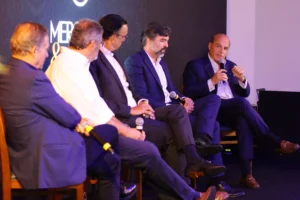 Cinco homens de terno participam de painel de debates no evento Mercado & Opinião, com um deles falando ao microfone.