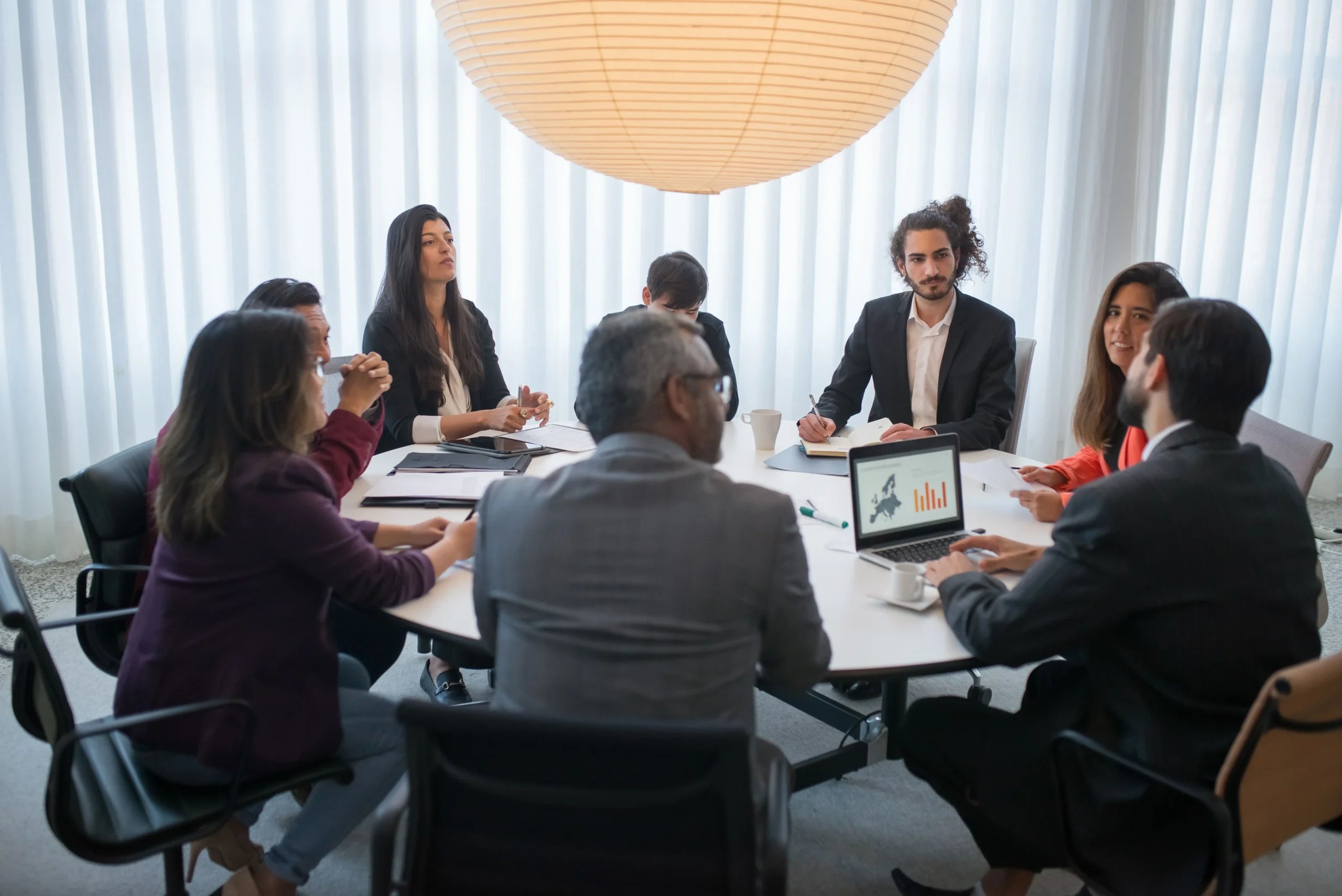 Grupo de profissionais de negócios reunidos ao redor de uma mesa redonda em sala de reunião, analisando documentos e gráficos em um laptop, sob luminária esférica de papel.