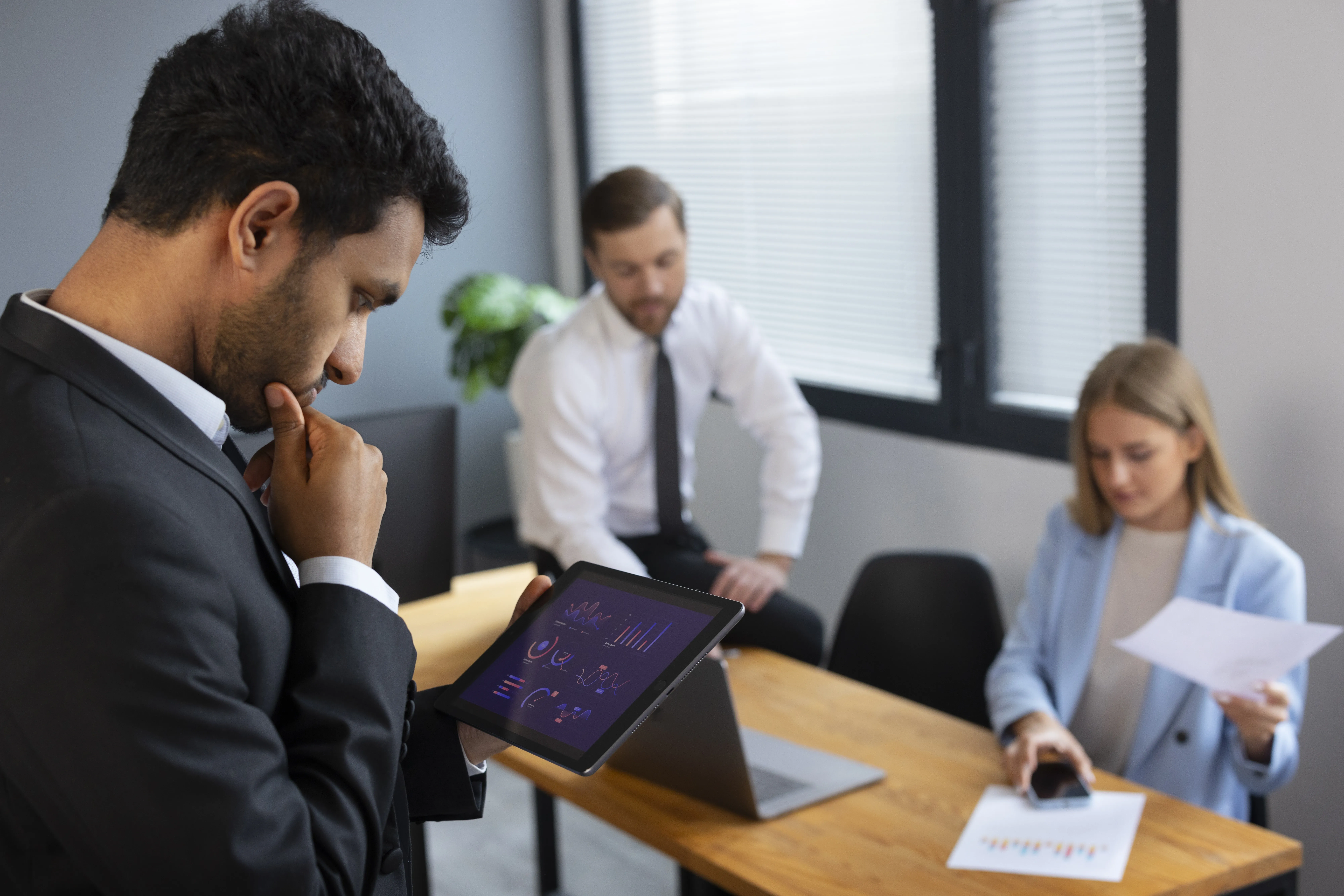 Equipe de negócios analisa dados financeiros em reunião enquanto homem de terno examina gráficos no tablet em primeiro plano.
