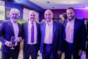 Quatro homens de terno posam sorrindo lado a lado em evento corporativo, um deles segurando uma taça de vinho, com iluminação azul e roxa ao fundo. Quatro homens de terno posam sorrindo lado a lado em evento corporativo, um deles segurando uma taça de vinho, com iluminação azul e roxa ao fundo. Quatro homens de terno posam sorrindo lado a lado em evento corporativo, um deles segurando uma taça de vinho, com iluminação azul e roxa ao fundo.