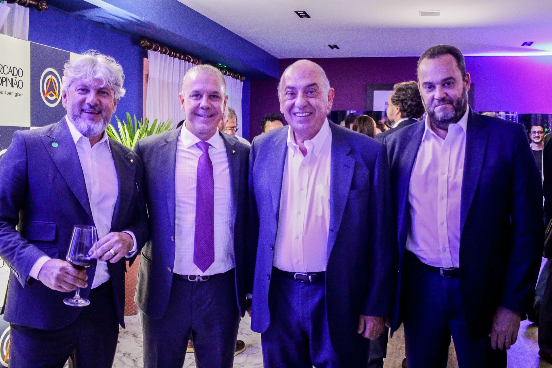 Quatro homens de terno posam sorrindo lado a lado em evento corporativo, um deles segurando uma taça de vinho, com iluminação azul e roxa ao fundo. Quatro homens de terno posam sorrindo lado a lado em evento corporativo, um deles segurando uma taça de vinho, com iluminação azul e roxa ao fundo. Quatro homens de terno posam sorrindo lado a lado em evento corporativo, um deles segurando uma taça de vinho, com iluminação azul e roxa ao fundo.