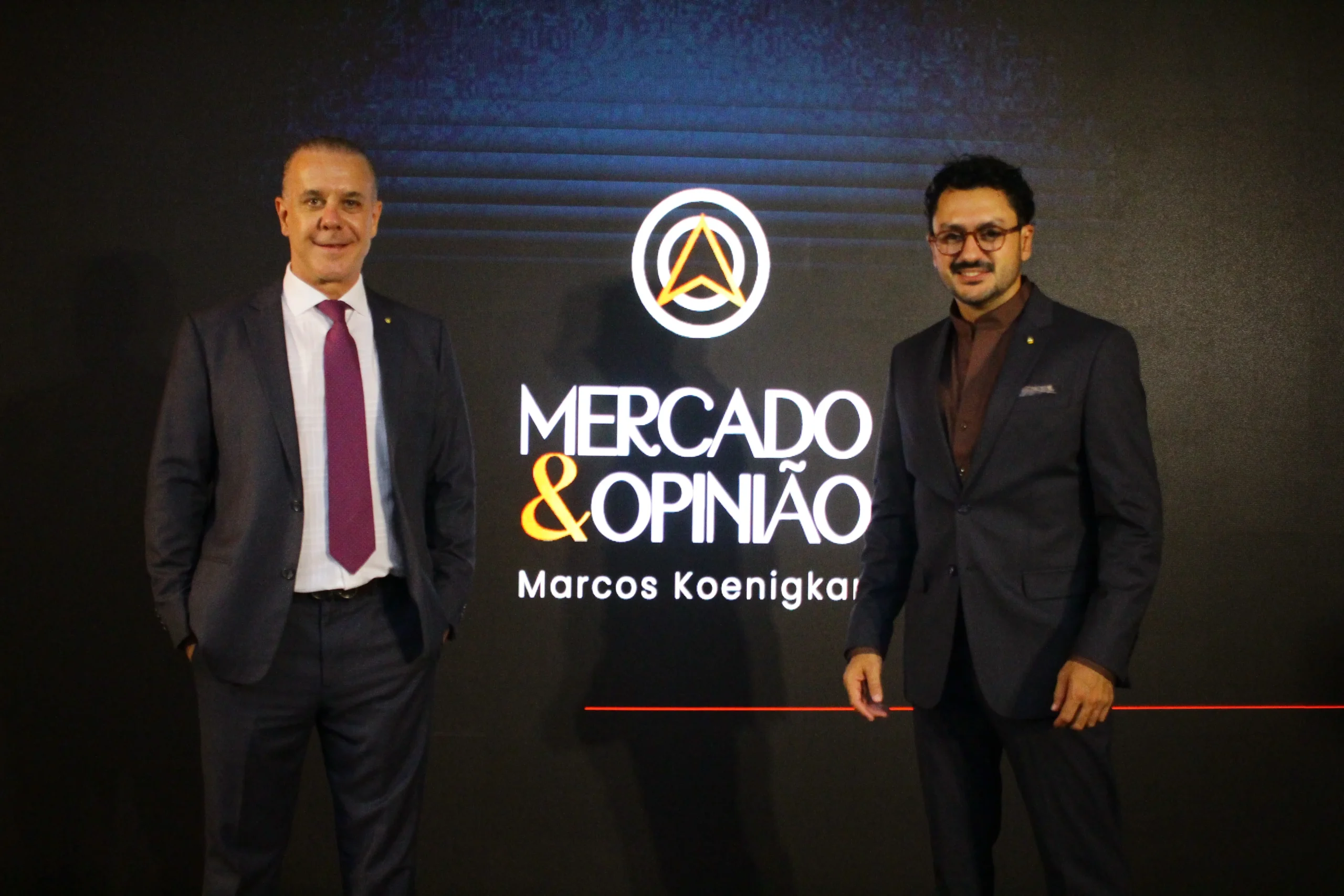 Dois homens de terno posam diante do painel do programa Mercado & Opinião, de Marcos Koenigkam.